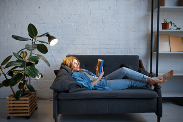 Young woman lying on cozy couch and reading a book