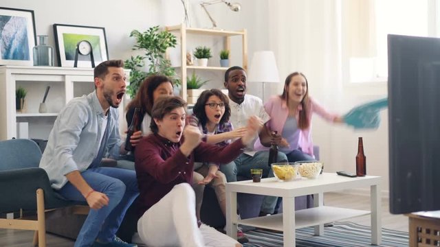 Excited girls and guys are cheering and hugging watching sports game on TV in apartment together. Modern active lifestyle, happy young and house concept.