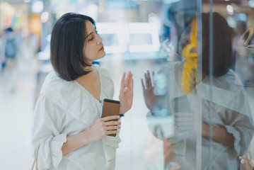 Young Asian beautiful woman shot hair look at window when shopping in a shopping mall. Woman standing and holding a shopping bag in the store near big glass window.