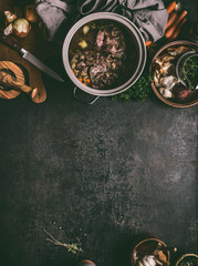 Food background with stewed beef meat in cooking pot on dark table with herbs, spices and kitchen utensils, top view, copy space. Country's meat dishes. Slow cooking. Rustic food