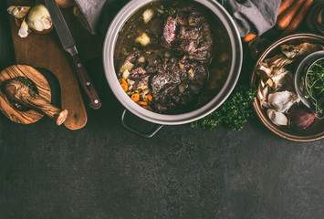 Stewed beef meat in cooking pot on dark rustic table with herbs, spices and kitchen utensils, top view, copy space. Country's meat dishes. Slow cooking