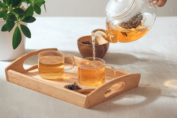 A hand pouring tea from glass teapot on wooden serving tray - obrazy, fototapety, plakaty