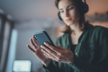 Young handsome girl relaxing at modern home,wearing casual clothes,playing music on smartphone and listening audio books in wireless headphones.Blurred background