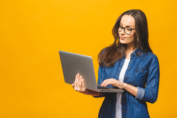 Young happy smiling woman in casual clothes holding laptop and sending email to her best friend. Isolated against yellow background. - obrazy, fototapety, plakaty