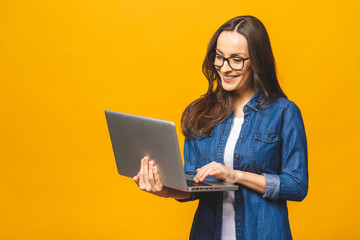 Portrait of a cheerful young woman wearing casual standing isolated over yellow background, holding laptop. - obrazy, fototapety, plakaty