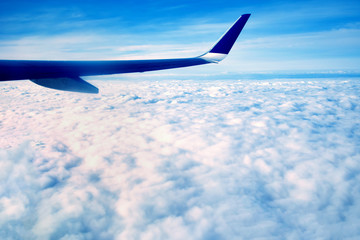 The blue wing of a large airplane, flying over the white morning clouds, at high altitude above the ground, against the blue sky on the horizon.