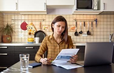 Thoughtful stressed young female sitting at kitchen table with papers and laptop computer trying to work through pile of bills, frustrated by amount of domestic expenses while doing family budget 