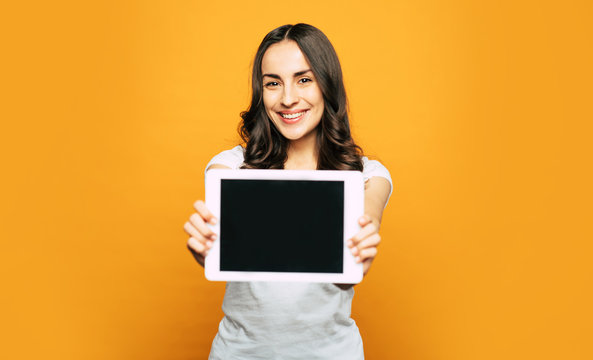 Come And See. A Tablet PC In The Hands Of Bodacious Smiley Girl With Slightly Curled Brownish Hair, Hazel Eyes And Stunning Smile In Front Of Spicy-orange Background.