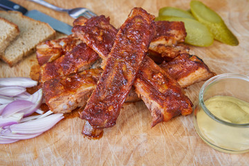 Close up Picture of american style bbq grilled pork spare ribs marinated in honey and served on wooden plate with slices of rustic bread, pickled cucumbers, chopped onions and french style hot mustard