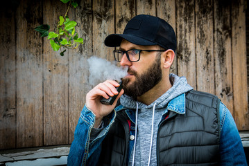 man with a beard smokes an electronic cigarette near wood wall