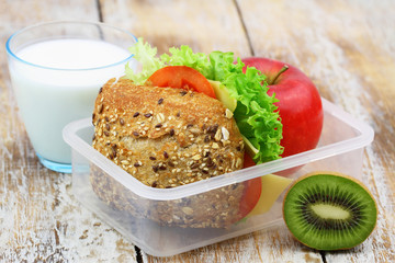 Nutritious lunchbox containing brown cheese roll with lettuce and tomato, apple, kiwi fruit and glass of milk