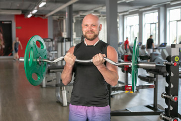 Fototapeta na wymiar Bald athlete in a black T-shirt and blue shorts with headphones, lifting the barbell with effort. He is standing in the gym and smiling