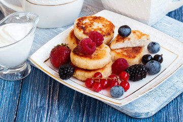 Russian syrniki or sirniki, cottage cheese fritters or pancakes served with raspberry, strawberry, blueberry, BlackBerry and cup of tea. Cottage cheese pancakes with fresh berries, sour cream and tea.