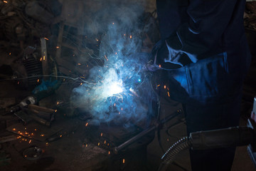 Close up of a male welder in blue uniform, welding mask and welders leathers, weld metal with an arc welding machine at the construction site, blue sparks fly to the sides