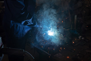 Close up of a male welder in blue uniform, welding mask and welders leathers, weld metal with an...