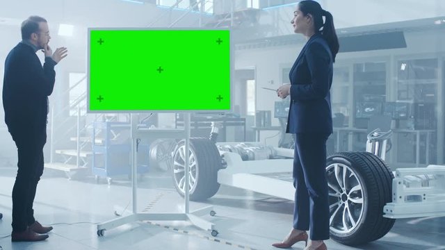 Male and Female Design Engineers are Discussing Something on a Interactive Whiteboard with Green Screen Next to an Electric Car Chassis Prototype. In High Tech Laboratory Facility with Vehicle Frame.
