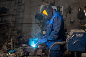 Strong man  is welding a metal construction in garage wearing mask, proctive glasses and blue uniform. Blue sparks are flying apart.