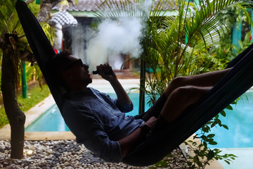 Man resting in a hammock and vaping and letting off steam from an electronic cigarette