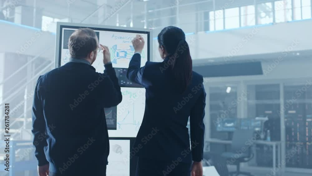 Poster Male and Female Design Engineers are Working and Discussing Something on a Interactive Whiteboard Next to an Electric Car Chassis Prototype. In High Tech Laboratory Facility with Vehicle Frame.