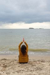 Woman practicing yoga against the blue sky and the azure sea on the sandy shore. Woman raises her arms to the sky in namaste posture and sits in a lotus pose.