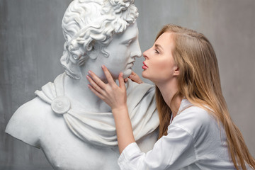 Young woman kissing God Apollo bust sculpture. Ancient Greek god of Sun and Poetry Plaster copy of a marble statue on grange concrete wall background. Ancient art and living beauty