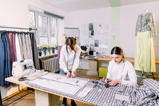 Two pretty women dressmakers working in the fashion atelier with stylish fabric on which they have marked out the pattern with tailors chalk. Checkered fabric, centimeter tape, tailoring scissors