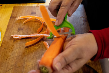 In the photo you can see the hands of a grandmother peeling carrots with a two-blade peeler. the color of the handle of the peeler is green. under the carrots is a yellowish wooden board with burns an