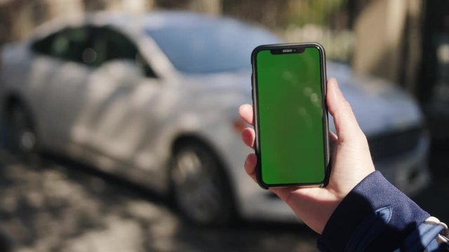 Close up view of male hand holding vertically modern smartphone with chromakey green screen on blurred white luxurious class car background. Successful lifestyle, rich accessories