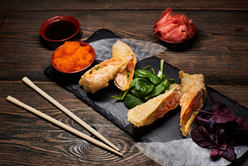 Asian dish serving with green, red basil, rice paper on stone plate. Sauces and bamboo chopsticks on wooden table. Salmon roasted in rice paper. Tempura recipe. Traditional Japanese cuisine. Top view.