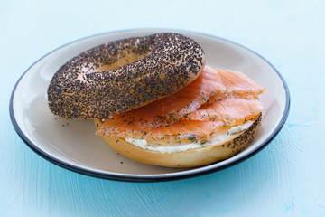 Poppy seeds bagel with cream cheese and smoked salmon served on a white plate with black rim. Blue table, high resolution