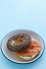 Poppy seeds bagel with cream cheese and smoked salmon served on a white plate with black rim. Blue table, high resolution