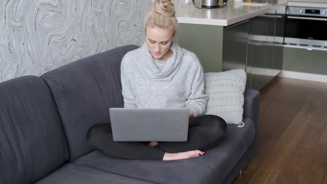 Cheerful young blond woman sitting on couch in living room and using laptop