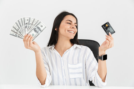 Photo of caucasian businesswoman 30s holding plastic credit card and fan with dollar money banknotes while sitting in armchair in office