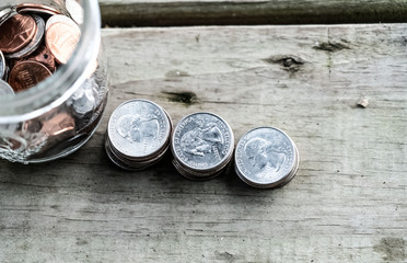 Focus on 3 stacks of quarters on old wooden floor board right next to a jar of various coins.