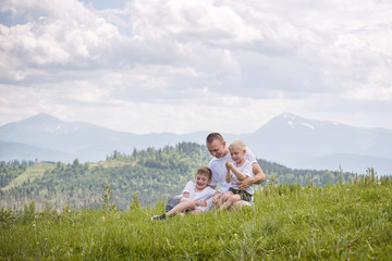 Fototapeta na wymiar Happy father with his two young sons sitting on the grass on a background of green forest, mountains and sky with clouds. Friendship concept.