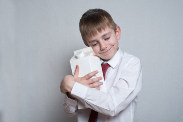 Little blond boy hugs a big white carton package. White shirt and red tie. Light background