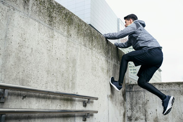 Stylish sportsman use architecture of urban area for his workout. He climbs at concrete wall, in the background modern office buildings.