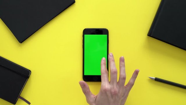 Woman sits at a yellow office Desk surrounded by office notebooks, folders and flips through the tape on her phone. Young woman leafing through the tape on her phone. prints text on your smartphone