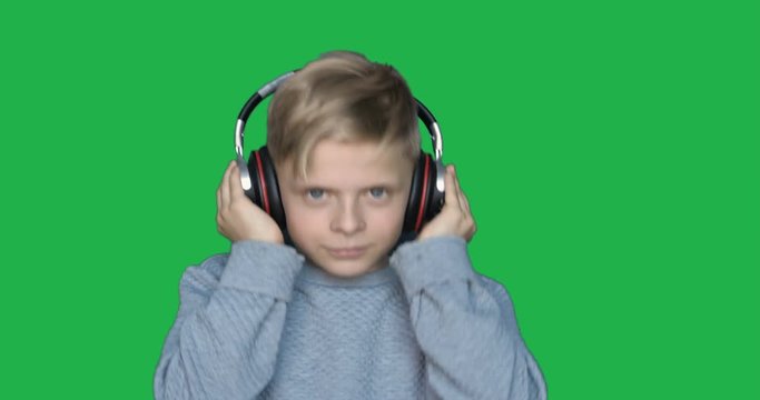 In slow-motion on the background of a chromakey, a young boy (boy) listens to music in wireless headphones and dances: Green background, Chromakey, BT headphones, Fashion, Blond, Music.
