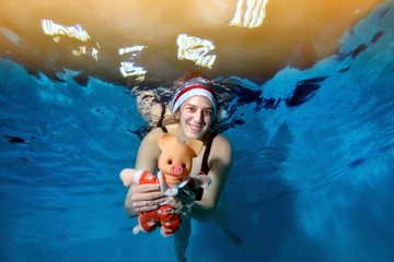 Obraz na płótnie Canvas A woman swims under water and holds a toy pig - a symbol of the New year. She poses for the camera with her eyes open and smiles with Santa's hat on her head. Portrait
