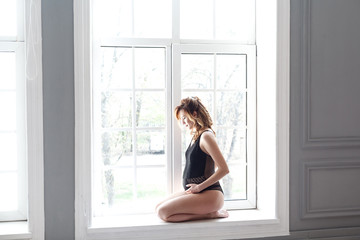 Side view portrait of beautiful pregnant young woman in black bodysuit sitting in front of window at home and holding with love her belly. Last months of pregnancy. Studio shot, light background,
