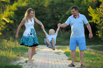 Naklejka na ściany i meble Mom and dad swing the little boy in their arms like a swing, and walk along the path in the Park at the sunset of a summer day. The concept of family happiness. Close up. Horizontal view