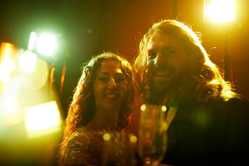 Jolly beautiful young couple with long curly hair standing in lights and smiling at camera while spending time at party