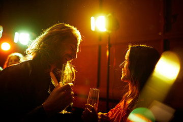 Cheerful handsome hipster man with long hair drinking champagne from flute and talking to pretty...