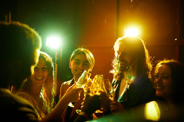 Group of hilarious young friends standing in circle and celebrating each other while drinking champagne at party in nightclub, disco lights effect