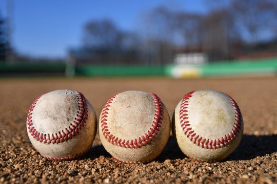 Photo of three game used baseballs on a baseball infield on a sunny day with a baseball field in the background
