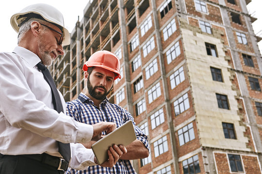 Discussion of the work plan. Senior professional engineer in formal wear and white helmet is showing project plan on digital tablet to young builder while standing at construction site.