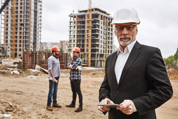 Visiting construction site. Portrait of confident elderly construction engineer in classic suit and hardhat is holding digital tablet and looking at camera while visiting building object