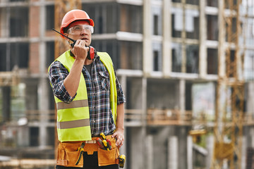 Сhecking work. Professional young builder in working uniform and red protective helmet talking to...