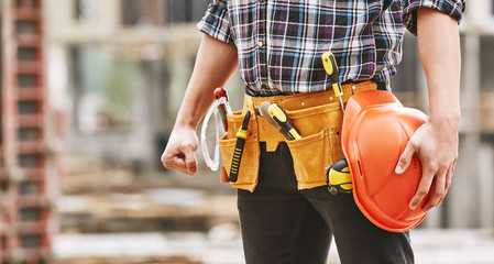 Fototapeta na wymiar Safe work. Cropped photo of male professional builder with construction tools holding a safety red helmet while standing outdoor of construction site